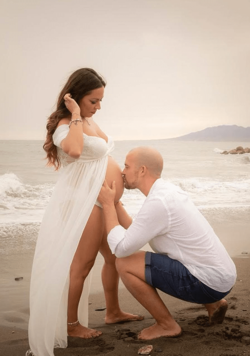 pareja en sesión de embarazo en la playa donde el padre besa la barriga de la futura mamá